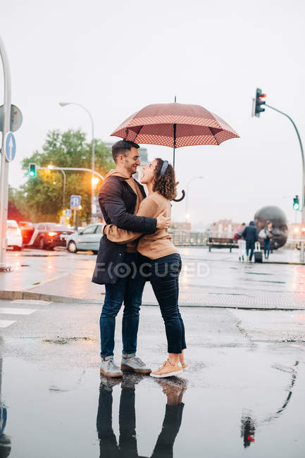 Side view of cheerful young man and woman with umbrella embracing and looking at each other while standing on street — Stock Photo
