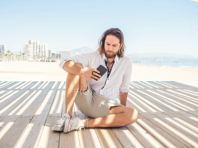 Bearded male browsing smartphone while sitting in gazebo on sandy beach near sea on sunny day — Stock Photo