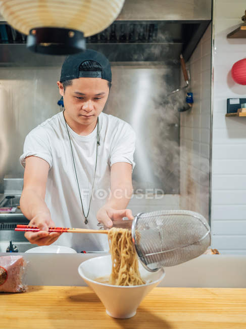 Giovane uomo mettendo tagliatelle calde in ciotola con le bacchette durante la cottura piatto giapponese in cucina — Foto stock