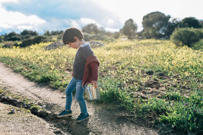 Чарівний маленький хлопчик, що носить невелике металеве відро під час прогулянки по дорозі в сонячній місцевості — стокове фото