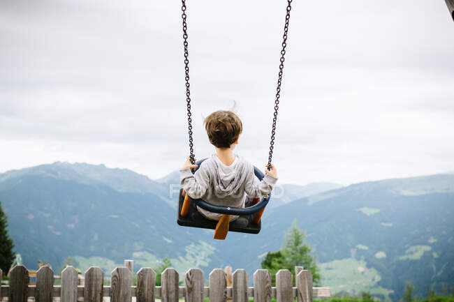 Visão traseira da criança ativa balançando alto no campo no fundo de montanhas de tirar o fôlego no dia nublado — Fotografia de Stock