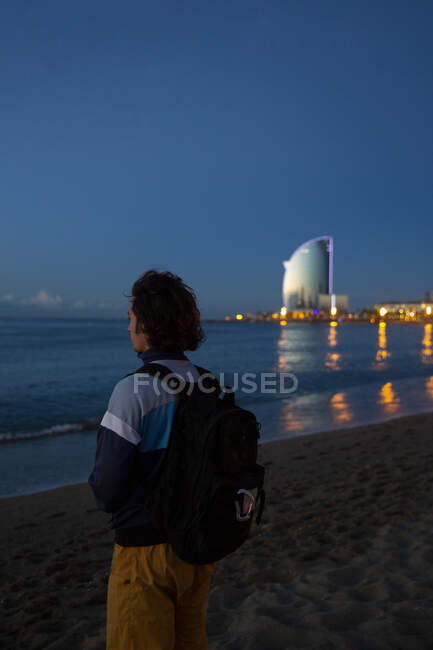 Back view of young man with backpack standing on sandy beach and looking along see and evening city — Stock Photo