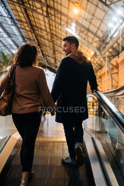 Vista posteriore di giovane uomo e donna che si tengono per mano mentre in piedi sulla passerella in movimento durante la data in un centro commerciale luminosamente illuminato — Foto stock