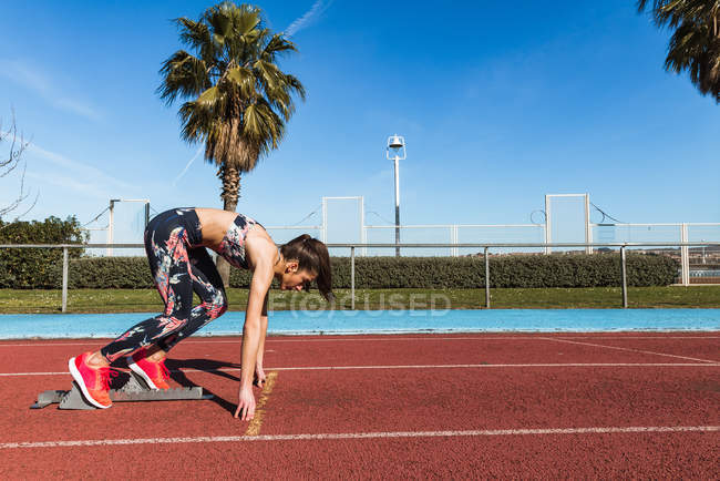 Schlanke junge Frau in Sportkleidung, die Füße im Startblock anlegt und die Strecke vor dem Lauf im Stadion berührt — Stockfoto