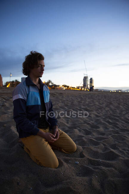 Сильный молодой человек с закрытыми глазами и сложенными вместе руками сидит на коленях на берегу моря на фоне заката — стоковое фото