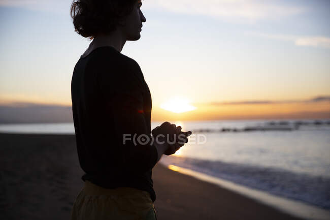 Vista lateral del hombre concentrándose en pensamientos con los ojos cerrados y las manos en gesto de oración de pie sobre las rodillas en la playa de arena a la luz del sol - foto de stock