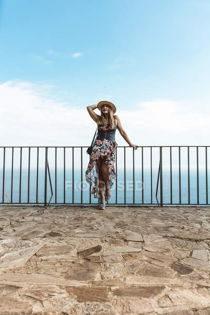 Женщина в летнем платье и соломенной шляпе, стоящая у забора на каменной террасе с морской накидкой на заднем плане — стоковое фото