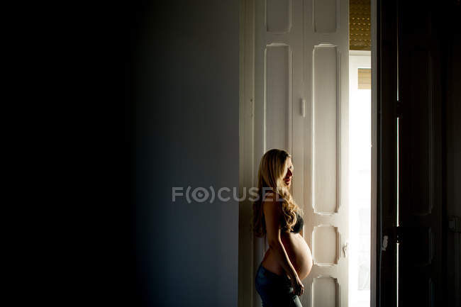 Вдумчивая беременная женщина в лифчике стоит у открытой двери дома — стоковое фото