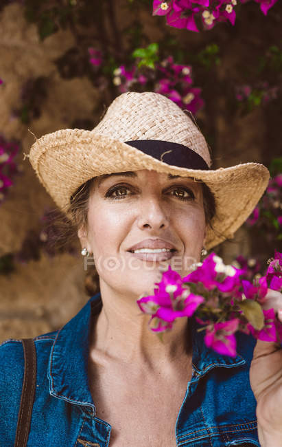 Frau mit Hut blickt in die Kamera, während sie in der Nähe einer Mauer mit rosa Blumen steht — Stockfoto