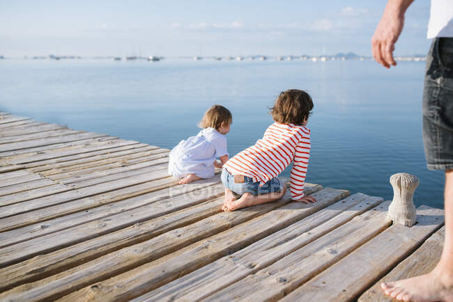 Crop man and adorable curious boy with toddler sister sitting on pier observing nature — Stock Photo