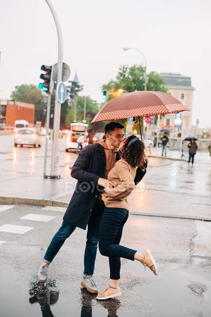 Side view of cheerful young man and woman with umbrella embracing and looking at each other while standing on wet city street on rainy day — Stock Photo