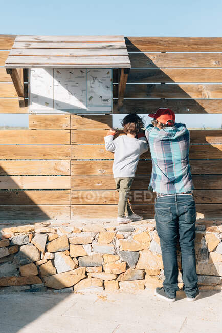 Visão traseira do pai com o menino olhando através do buraco na cerca de madeira observando a natureza — Fotografia de Stock