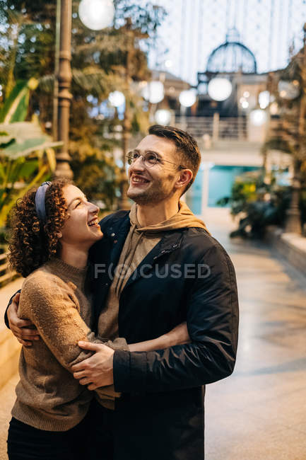 Cheerful young man and woman embracing and looking at each other while standing inside illuminated pavilion during date — Stock Photo
