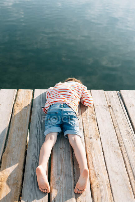 Desde arriba de niño frío acostado con la cara hacia abajo en el muelle de madera en el sol con agua pacífica por debajo - foto de stock
