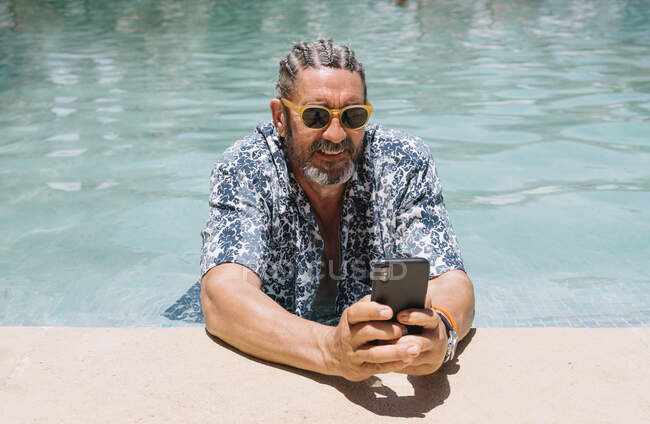 Barbudo macho maduro en elegantes tonos y camisa de navegación smartphone mientras se relaja en agua limpia de la piscina - foto de stock