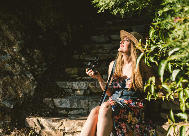 Путешествующая женщина в платье и шляпе держит камеру, сидя на каменных ступеньках на солнечной улице — стоковое фото
