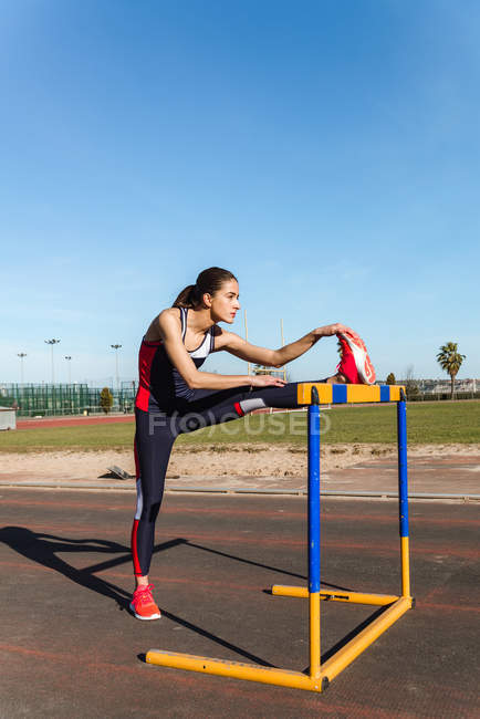 Jeune femme forte en vêtements de sport s'étirant sur l'obstacle contre le ciel bleu pendant l'entraînement sur le stade — Photo de stock