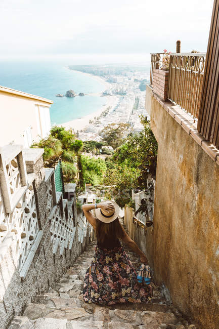 Вид ззаду на невпізнавану жінку в літньому вбранні, що стоїть на вулиці кам'яні сходи з морським узбережжям на фоні — стокове фото