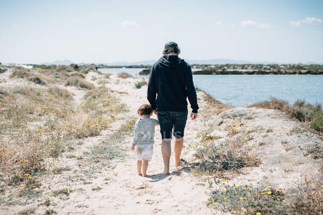 Вид сзади на молодого человека в темной одежде, держащегося за руки с ребенком и гуляющего по песчаному берегу с сухой зеленью в солнечный день — стоковое фото