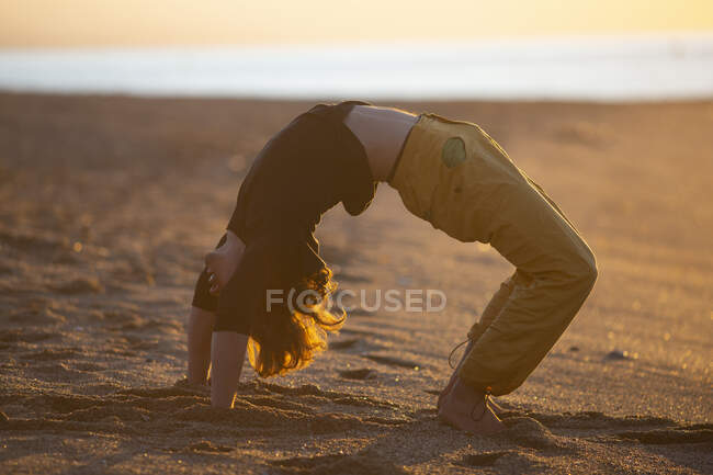 Sportive man in yellow pants barefoot performing yoga meditation workout on sand beach in sunset evening — Stock Photo