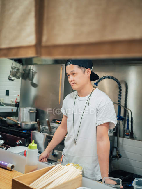 Молодой человек в белой футболке и черной шапке готовит рамен в японском ресторане — стоковое фото