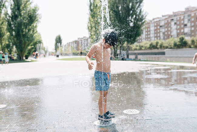 Little boy in swimsuits standing near jet of water splashing out of fountain on street — Stock Photo