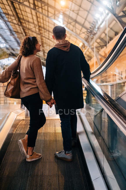 Back view of young man and woman holding hands while standing on moving walkway during date in brightly illuminated mall — Stock Photo