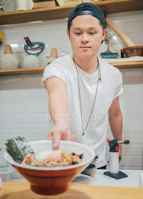 Jovem cozinheiro masculino colocando no balcão tigela grande com delicioso prato japonês chamado ramen no café oriental — Fotografia de Stock