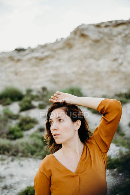 Porträt einer nachdenklichen Frau in Bluse, die die Hand über dem Kopf vor dem Hintergrund einer wilden Wüstenlandschaft hält — Stockfoto