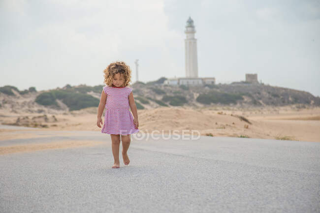 Кучеряве дитя в смугастій ходьбі на піщаному пляжі на розмитому фоні природи — стокове фото