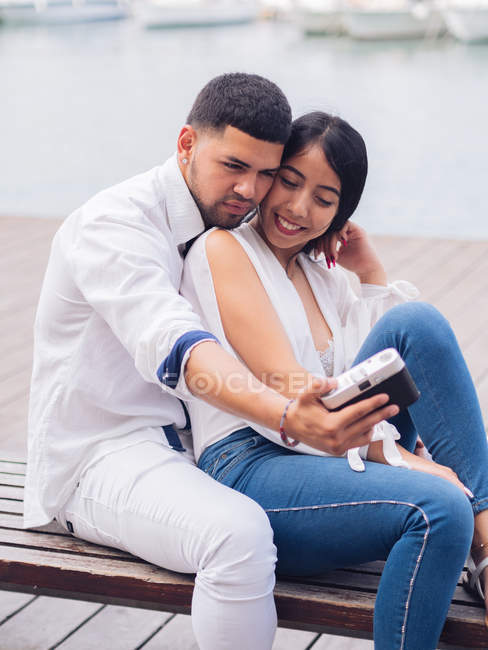 Young couple sitting and having fun taking a selfie picture on wooden bench on sandy seaside — Stock Photo