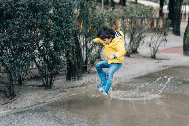Excited boy in coat and gumboots having fun on street and jumping on puddle — Stock Photo