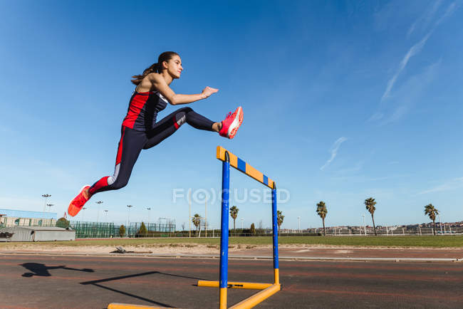 Strong young woman in sportswear leaping over hurdle against blue sky during workout on stadium — Stock Photo