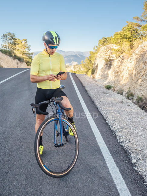 Homem saudável descansando e usando smartphone com bicicleta em uma estrada de montanha em um dia ensolarado — Fotografia de Stock