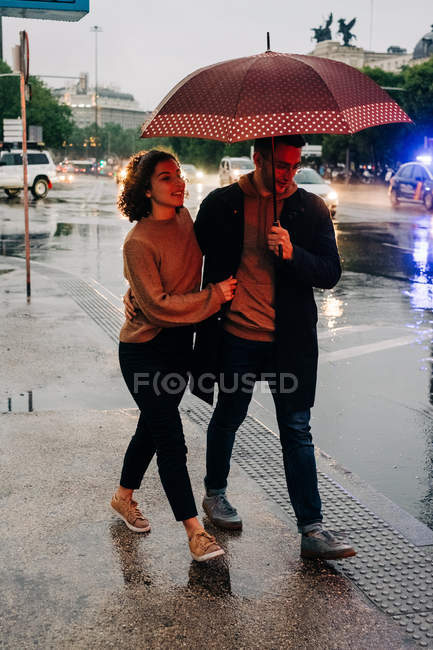 Side view of cheerful young man and woman with umbrella embracing while standing on wet city street on rainy day — Stock Photo