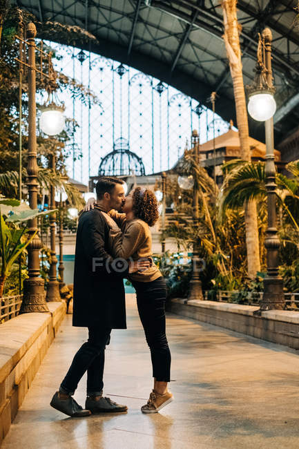 Cheerful young man and woman embracing and kissing looking at each other while standing inside illuminated pavilion during date — Stock Photo