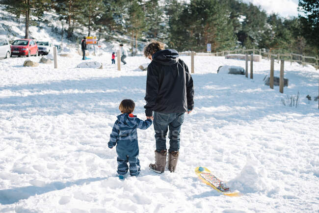 Back view of young man in dark warm clothes holding hands with kid in striped overalls and walking in snow in sunny day — Stock Photo