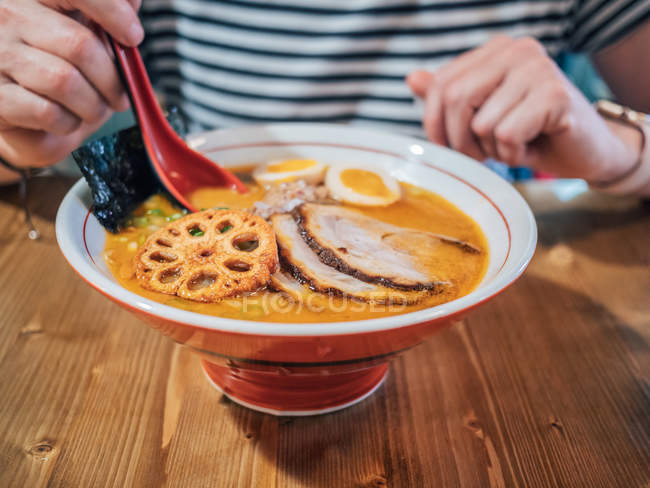 Cropped view of woman sitting at wooden table and enjoying bowl of delicious ramen topped with egg and sliced pork — Stock Photo