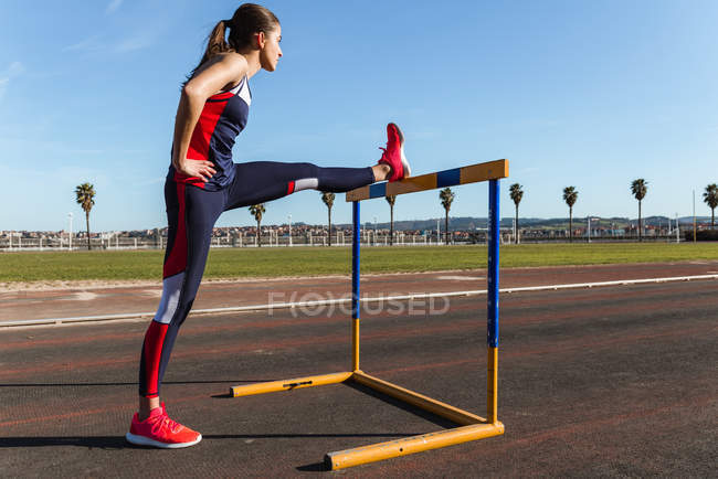 Jeune femme forte en vêtements de sport s'étirant sur l'obstacle contre le ciel bleu pendant l'entraînement sur le stade — Photo de stock