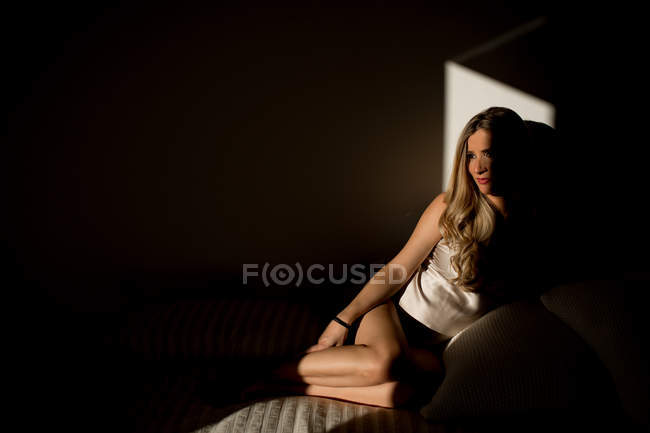 Элегантная беременная женщина смотрит в сторону, сидя на удобной кровати под солнечным светом в темной комнате дома — стоковое фото