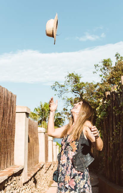 Aufgeregte Frau wirft Strohhut auf schmaler Straße in die Sonne — Stockfoto