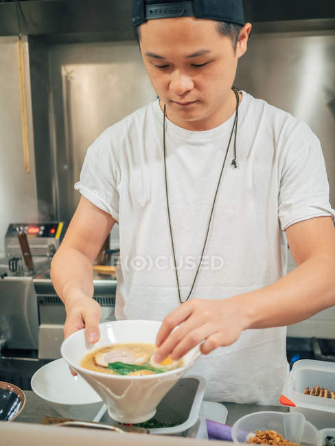 Asian man putting halved egg in bowl with fresh cooked ramen in restaurant kitchen — Stock Photo