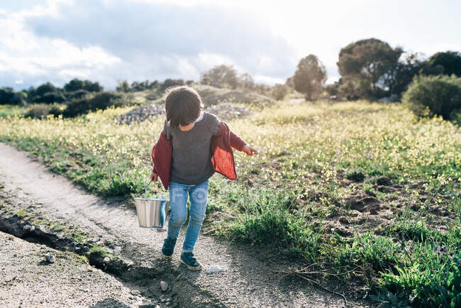 Adorable little boy carrying small metal bucket while walking on road in sunny countryside — Stock Photo