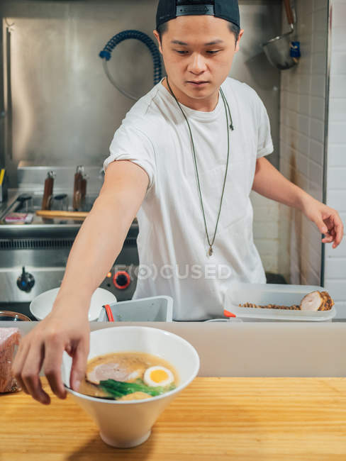 Young man putting bowls of fresh cooked traditional Japanese dish on wooden counter in restaurant — Stock Photo