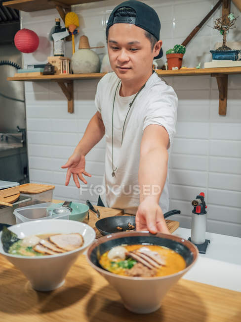 Jeune homme mettant des bols de plat japonais traditionnel cuit frais sur le comptoir en bois dans le restaurant — Photo de stock