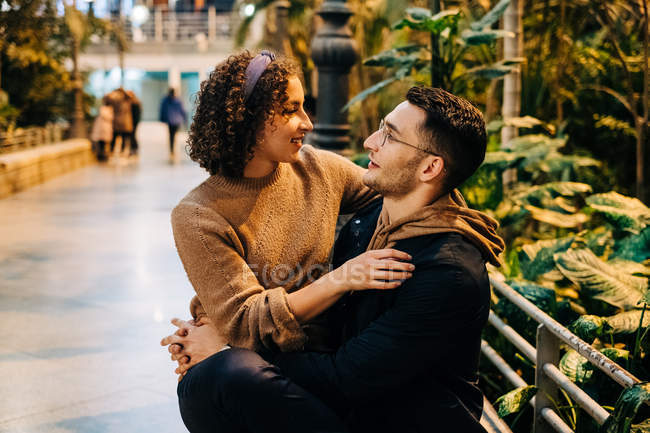Seitenansicht eines jungen Mannes und einer jungen Frau, die sich umarmen und einander anschauen, während sie bei einem romantischen Date auf der Stadtstraße auf einer Bank sitzen — Stockfoto