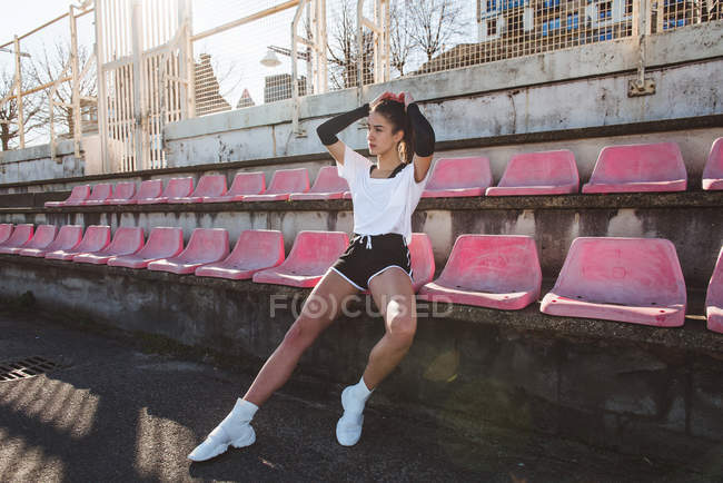 Schlanke junge Frau in Sportbekleidung schaut beim Training weg, während sie auf dem grunzenden Stadionsitz sitzt — Stockfoto