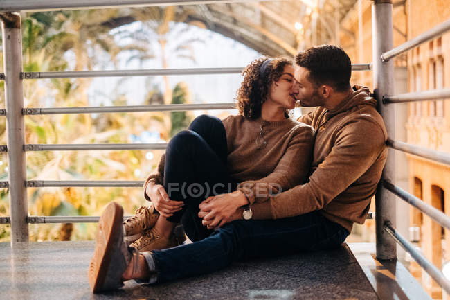 Cheerful young man and woman embracing and kissing looking at each other while sitting inside illuminated pavilion during date — Stock Photo