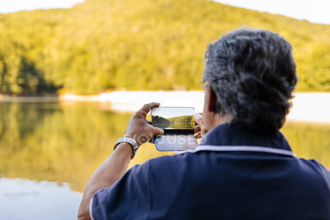 Visão traseira de um homem sênior tirando foto com telefone celular enquanto contempla paisagem incrível montanha lago — Fotografia de Stock