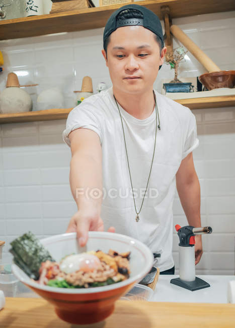 Young man standing in kitchen and handing bowl with fresh cooked Japanese dish — Stock Photo